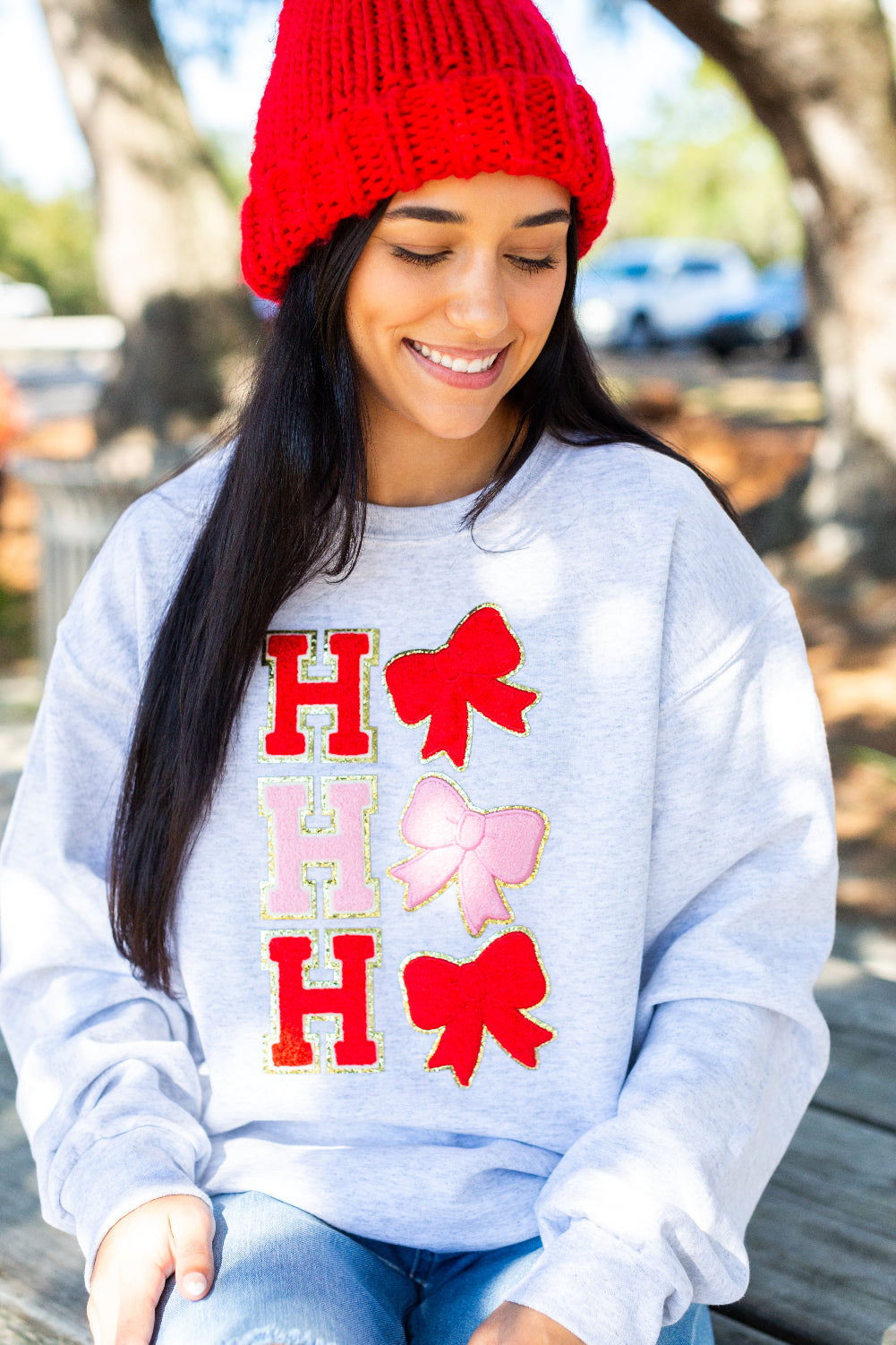 Woman wearing a light blue sweatshirt with red and pink bows and 'HO HO HO' text, sitting outdoors.
