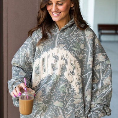 Person wearing a camouflage hoodie with 'COFFEE' text, holding a drink.