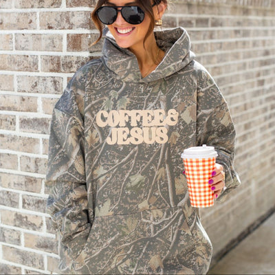 Person wearing a camouflage hoodie with 'COFFEE IS JESUS' text, holding a coffee cup against a brick wall.