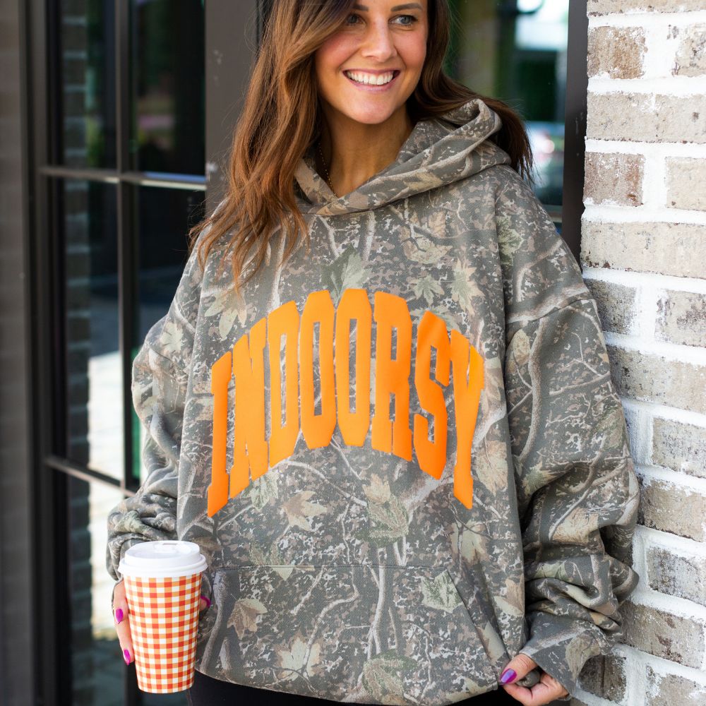 Woman wearing a camouflage hoodie with orange text, holding a coffee cup, standing against a brick wall.