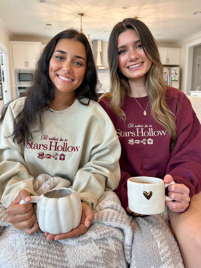 Two women sitting in a kitchen holding mugs with 'Stars Hollow' text. Gilmore girls sweatshirt