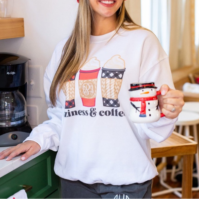 Woman wearing a white sweatshirt with graphics and text, holding a mug in a kitchen.