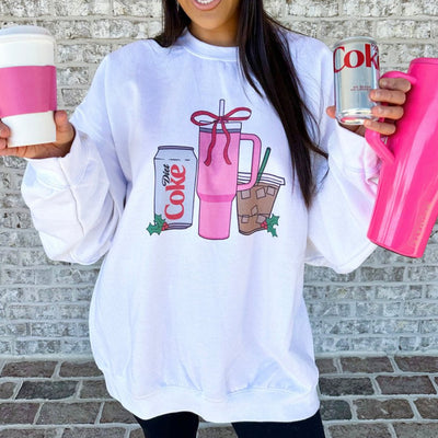 Person wearing a white sweatshirt with drink graphic, holding pink and white cup, can, and tumbler.